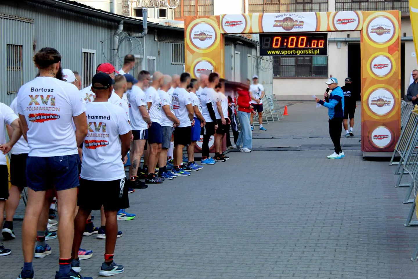 Zawodnicy w koszulkach XVI Półmaratonu ustawieni na starcie pod bramą z zegarem na dziedzińcu Zakładu Karnego w Rawiczu